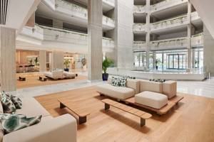Lobby des Hipotels Mediterraneo mit Sofas, Tischen und einem Swimmingpool im Hintergrund.