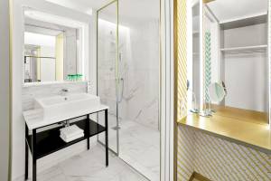 Das elegante Badezimmer des MAXX by Steigenberger Vienna mit weißen Marmorfliesen, goldenen Akzenten und einer modernen Dusche.