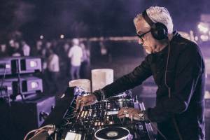 Ein älterer DJ mit weißer Frisur und schwarzer Kleidung spielt Musik in einer Nachtclub-Umgebung.
