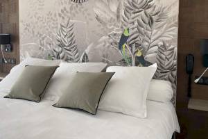 Ein Bett mit weißen Laken und grauen Kissen vor einer Wand mit exotischen Vogelmalereien im La Lagune Hotel.