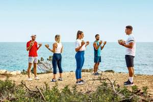 Gruppe von Menschen, die auf einem Strand Fitnessübungen mit Steinen machen, Victoria Menorca Hotel.