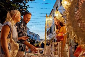 Drei Personen betrachten Schmuck auf einem Marktstand im Abendlicht von Victoria Menorca.
