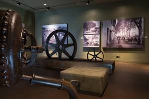 Museumsausstellung mit einem großen Rad und mehreren alten Schwarz-Weiß-Fotos an der Wand.