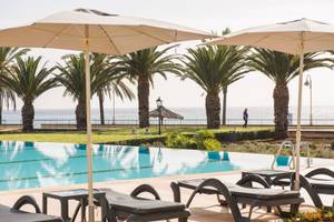 Außenpool des Vila Galé Santa Cruz mit Liegestühlen, Sonnenschirmen und Palmen am Meer