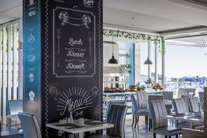 Innenbereich des Asterias Beach Resort Restaurants mit einem großen schwarzen Menüboard, das neben einem Tisch steht, mit Blick auf einen sonnigen Hafen.