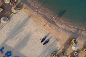 Luftaufnahme des Desert Rose Resorts Strands mit mehreren Liegen, Sonnenschirmen und blauen Booten im Sand.