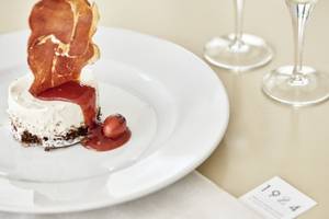 Ein Gericht auf einem weißen Teller serviert, dekoriert mit einem Stück Prosciutto, einer roten Soße und einer kleinen Tomate.