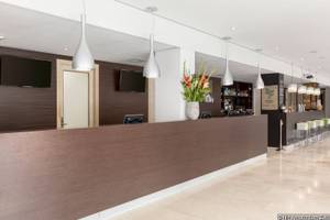 Innenansicht der Rezeption im NH Amsterdam Zuid Hotel mit Holztresen, Blumenvase, Monitoren und Barhockern.