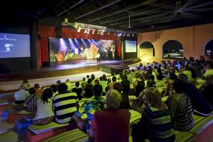 Ein Publikum sitzt in einem Theatersaal und schaut eine Live-Show auf der Bühne, während ein Mann in schwarzer Kleidung auf der Bühne steht, während es Nacht ist.
