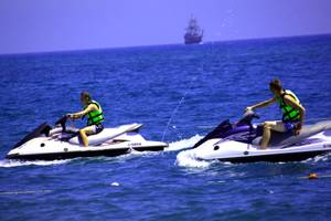Zwei Personen auf Jetskis im Meer, ein Segelschiff im Hintergrund.
