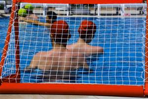 Schwimmer im Schwimmbad mit roten Hüten spielen Wasserball vor einem roten Tor.