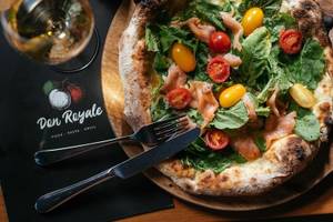 Eine Pizza mit Lachs und Tomaten auf einer Holzplatte mit Messer und Gabel.
