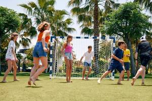 Mehrere Kinder spielen Fussball auf einem Kunstrasenplatz im Freien, vor einem Tor.