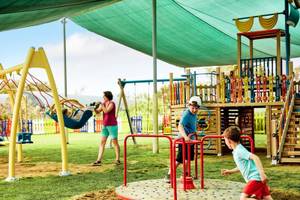 Familie auf dem Spielplatz im Magic Life Jacaranda Hotel, Kinder spielen auf Schaukeln und Karussell.