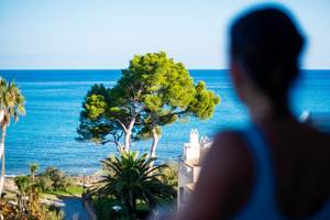 Frau blickt auf das Meer und einen Baum in der Ferne vom Balkon aus.