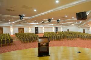 Saal im Alan Xafira Deluxe Resort & Spa mit vielen Stühlen, einem Podium und Lautsprechern an der Decke.