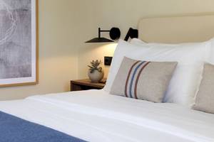 Bett im Domes Aulus Zante Hotel mit weißen Laken und Kissen, neben einem Nachttisch mit Pflanze.