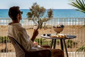 Ein Mann sitzt auf einem Balkon mit Blick auf das Meer und hält ein Glas Wein.