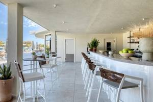 Innenansicht der Bar im Domes Aulus Zante Hotel mit weißen Möbeln, Tischen und Stühlen, Pflanzen und einer offenen Aussicht auf den Außenbereich.