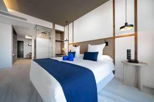Innenansicht eines Zimmers im H10 Costa Mogan Hotel mit einem Bett, blauen Kissen, weißen Decken und einer modernen Einrichtung.