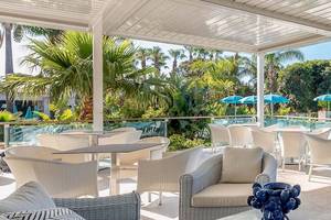 Terrasse im Acacia Palace Hotel mit weißen Möbeln, blauen Sonnenschirmen und Blick auf einen Pool und Palmen.