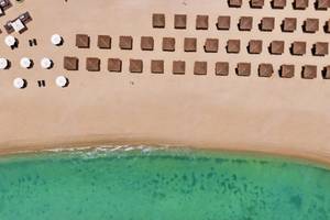 Luftaufnahme eines Strandes mit vielen braunen Strohhütten und weißen Sonnenschirmen.