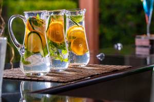 Drei Glaskruecke mit Wasser, Orangenscheiben, Gurkenscheiben und Minze auf einem Tisch.