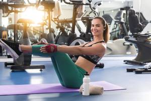 Frau im Fitnessstudio sitzt auf Matte und lächelt, während sie eine Beinübung macht