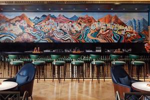 Innenansicht der Bar im St. Regis Al Mouj Muscat Resort mit buntem Wandgemälde und grünen Barhockern.