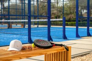 Tennisschläger, Ball, Hut und Sonnenbrille auf einer Holzbank neben einem Tennisplatz