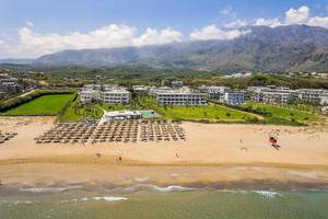 Luftaufnahme des Vantaris Palace Resorts am Strand mit vielen Sonnenschirmen und Bergen im Hintergrund.