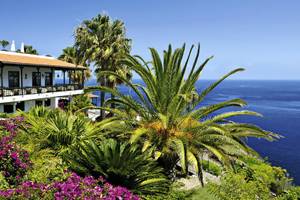 Außenansicht des Jardin Tecina Hotels mit Blick auf das Meer und eine Terrasse mit Palmen und Blumen.