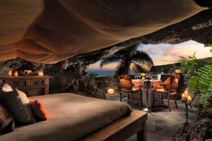 Ein Schlafzimmer, das in einen Felsen gehauen ist, mit Blick auf den Ozean und einem Esstisch mit Kerzen davor.