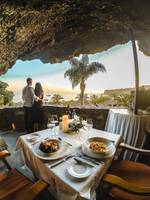 Ein Paar genießt ein Abendessen in einem Gartenrestaurant mit Meerblick, bei Sonnenuntergang.
