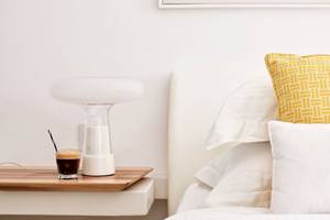 Hotelzimmer des Myconian Ambassador Relais & Chateaux mit Bett, gelbem Kissen und Nachttisch mit Lampe und Getränk.